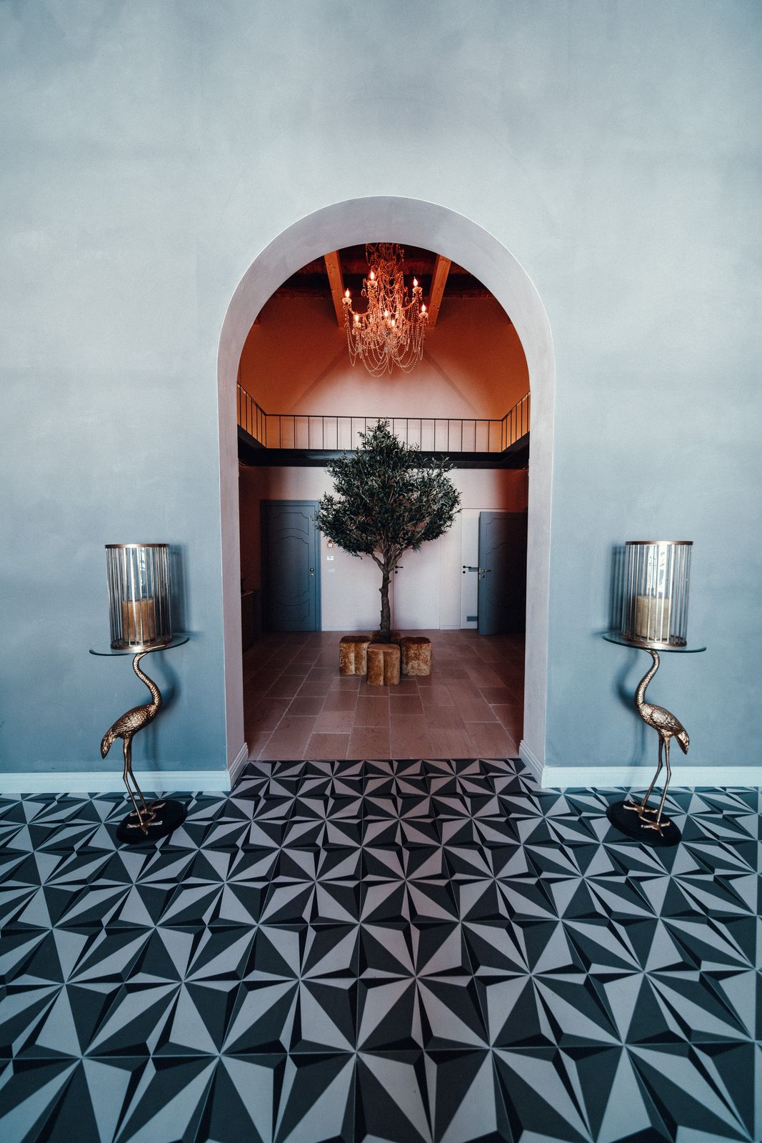 Brass crane candle stands either side of an arched doorway, framing an olive tree and a chandelier in the warm room beyond.