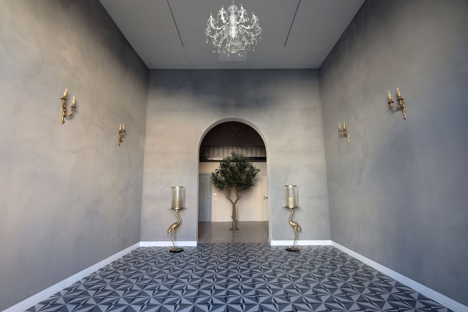 Grey plastered hall with a crystal chandelier, brass crane candle stands and a geometric tiled floor, opening through an arch to the olive tree beyond.