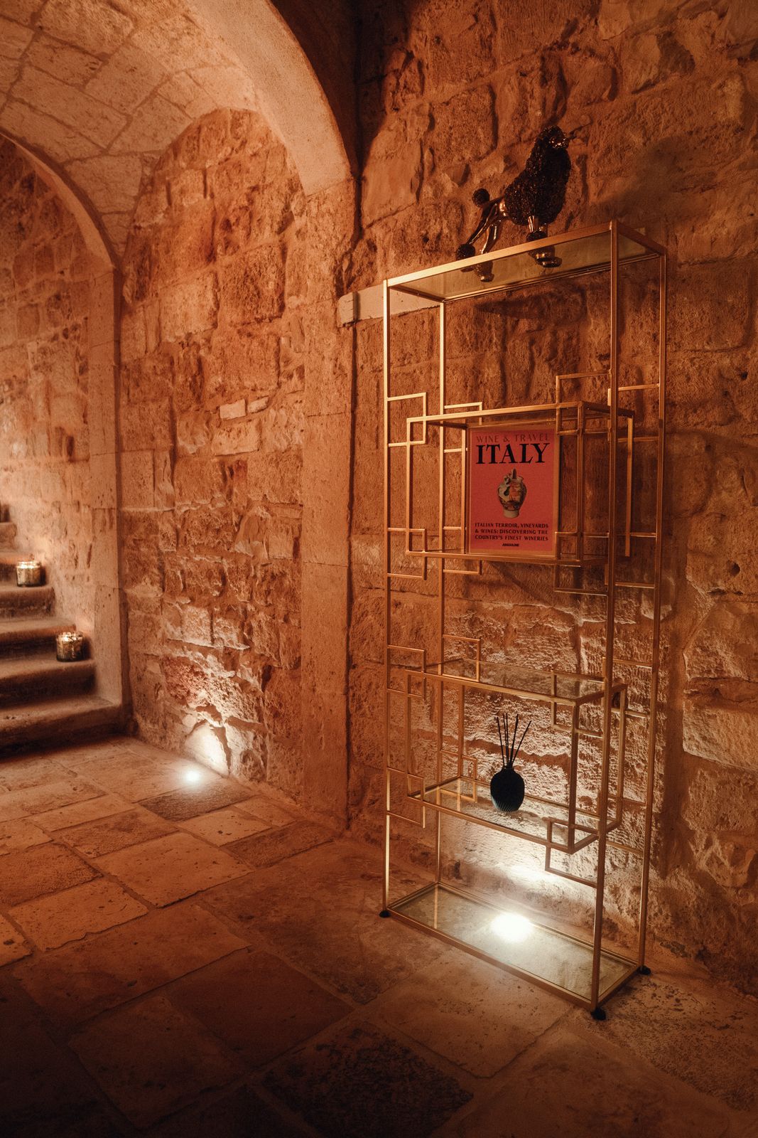 A slim gold shelf holding books and objects in a vaulted stone passage, with candlelit steps beyond.