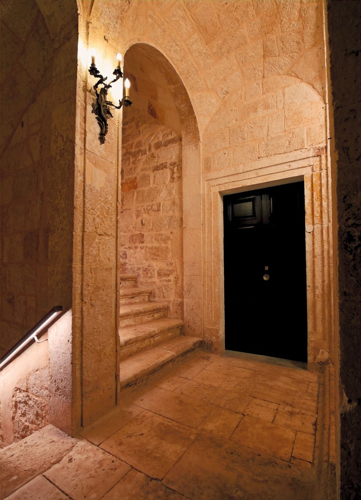 A lit sconce and the arched passage to a dark door.