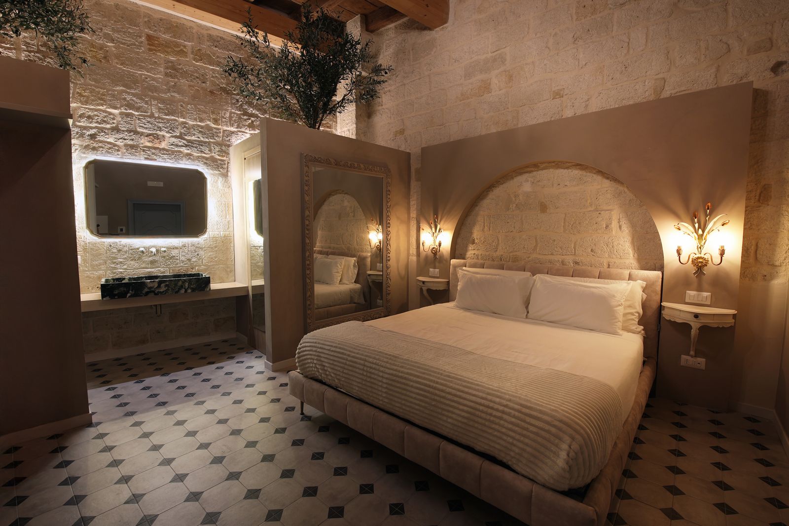 Garden Deluxe Room: a bed set into an arched stone alcove, with a green marble basin and an olive tree beneath the wooden beam ceiling.