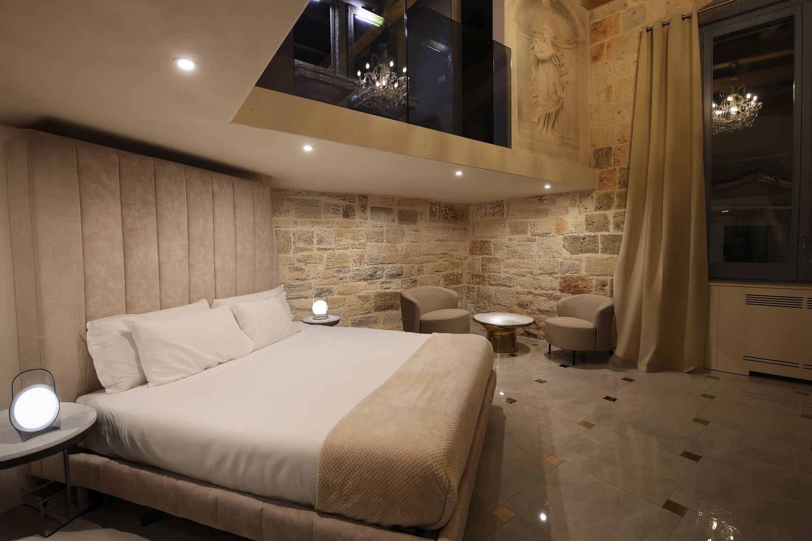 Twins Junior Suite: a velvet-headboard bed below a mezzanine with a glass balustrade, beside a carved figure set into the stone wall.