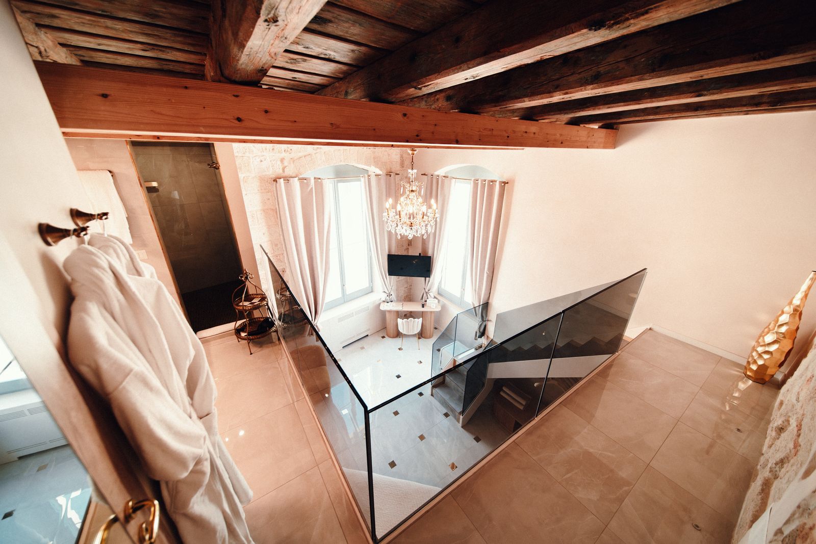 Twins Junior Suite: the view from the mezzanine, past a glass balustrade and the staircase, to the chandelier and curtained windows beneath the beamed ceiling.