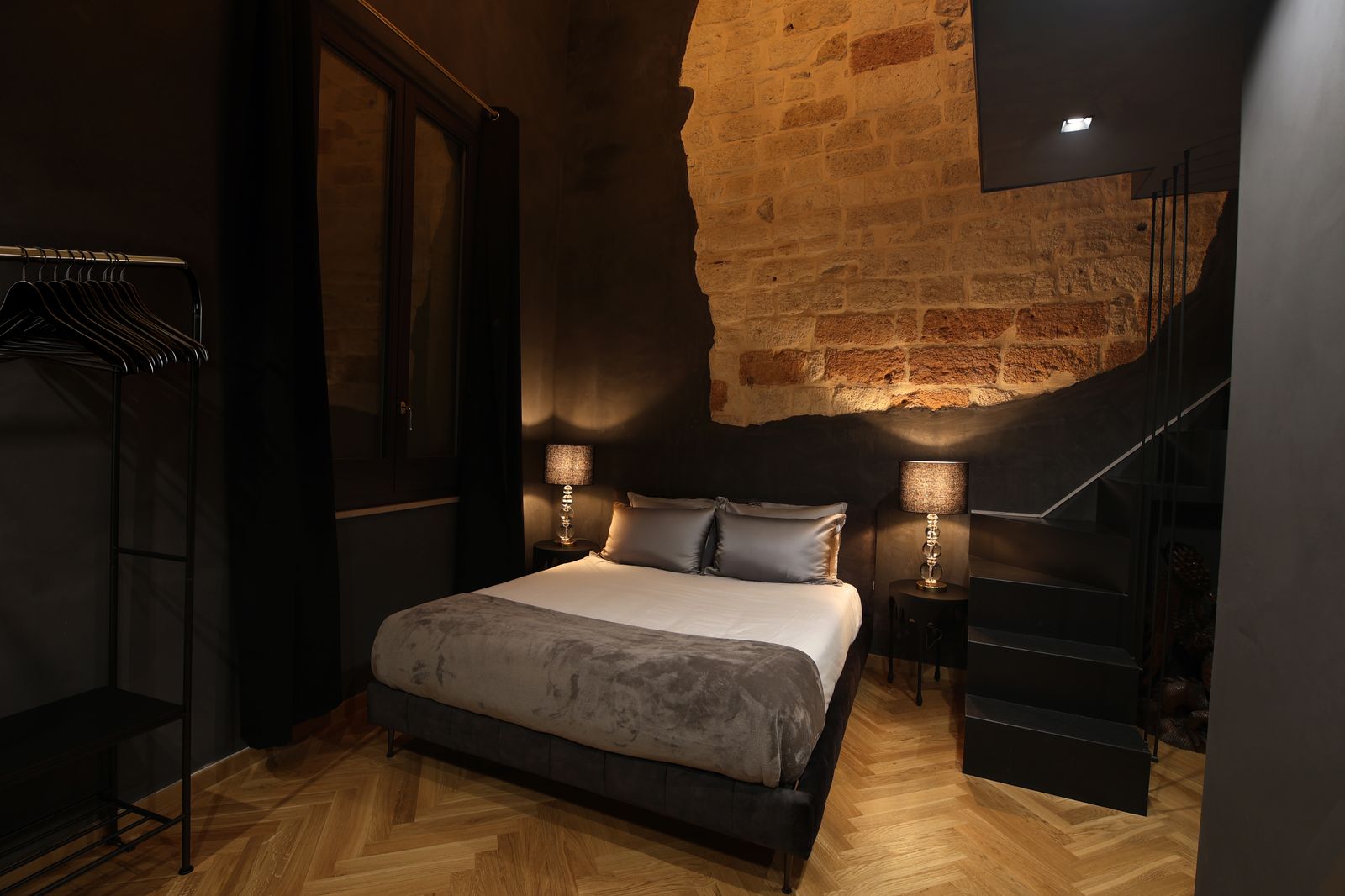 Firestone Junior Suite: dark plastered walls and herringbone parquet, with a lit patch of raw stone above the bed and stairs to the upper level.
