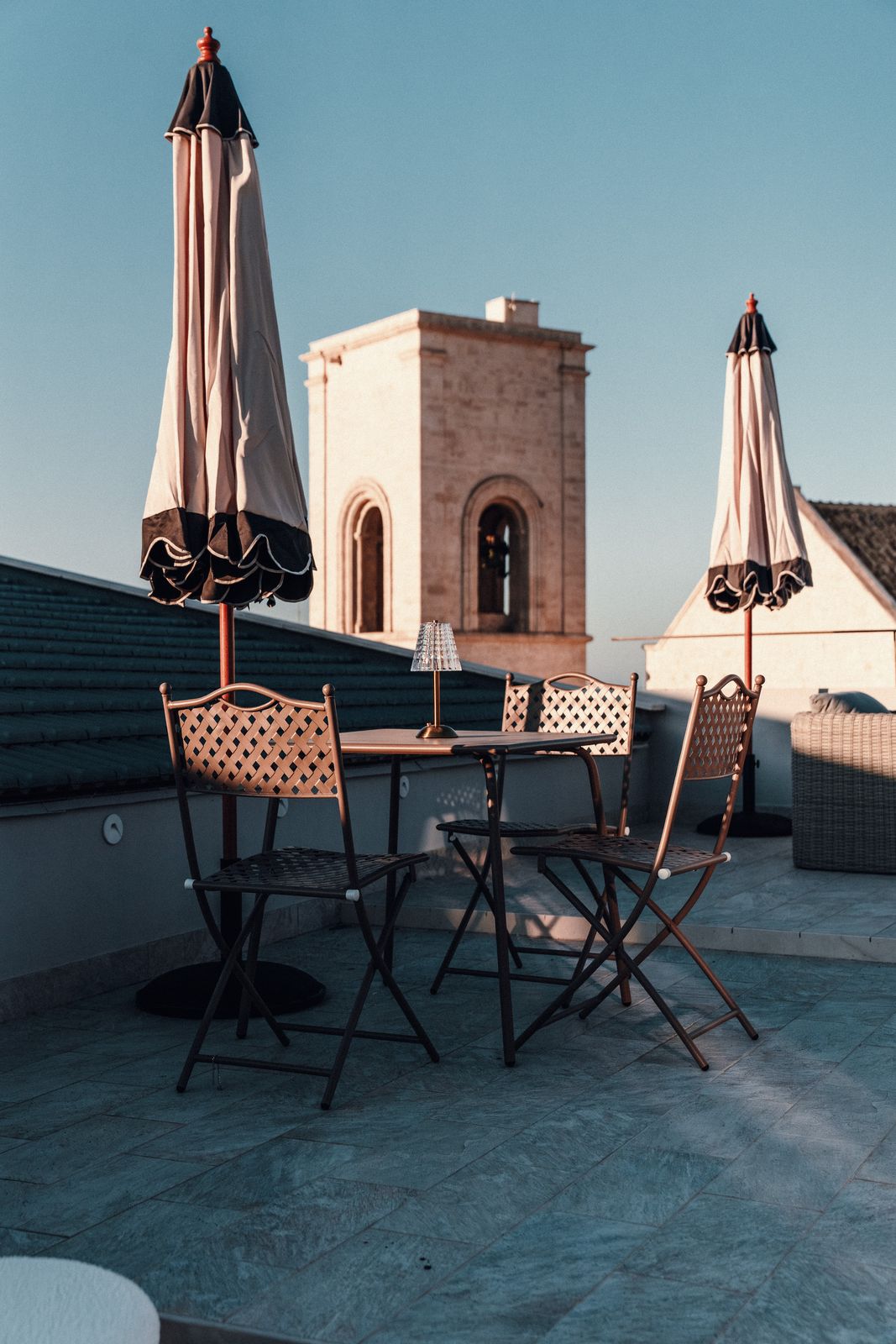 A bistro table and chairs set beneath parasols on the rooftop, facing Polignano's stone bell tower.
